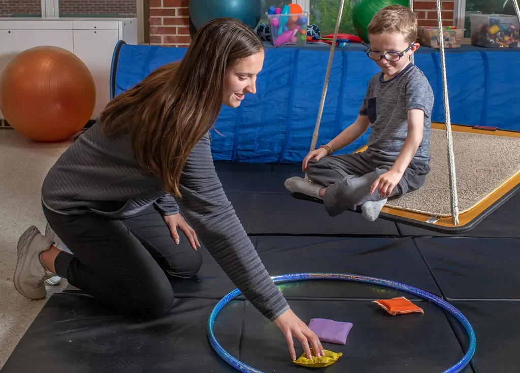 An M S O T student reaches down for a small bean bag to give to a child sitting on a swing